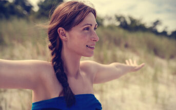 A woman with a braid stands outdoors with her arms outstretched, smiling and looking to the side. She is wrapped in a blue towel and has a background of tall grass and trees. The weather appears to be sunny.