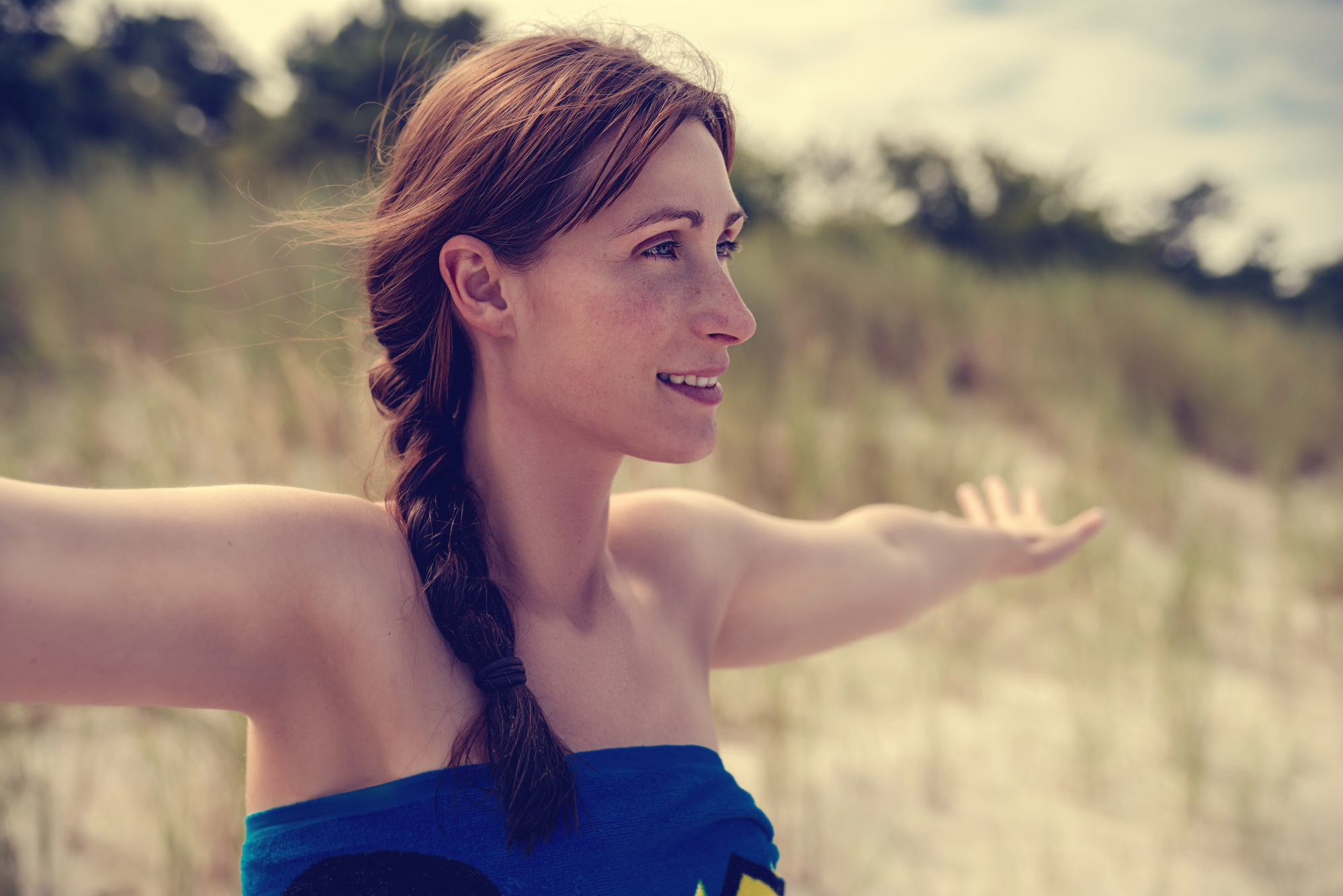 A woman with a braid stands outdoors with her arms outstretched, smiling and looking to the side. She is wrapped in a blue towel and has a background of tall grass and trees. The weather appears to be sunny.