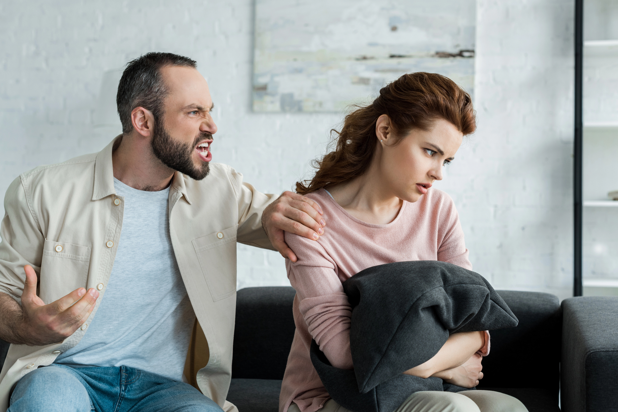 A man with a beard and bald spot angrily gestures and speaks to a woman with red hair who looks away, upset. She sits on a couch clutching a gray pillow, her body turned slightly away from him. The background features a white wall with abstract art.