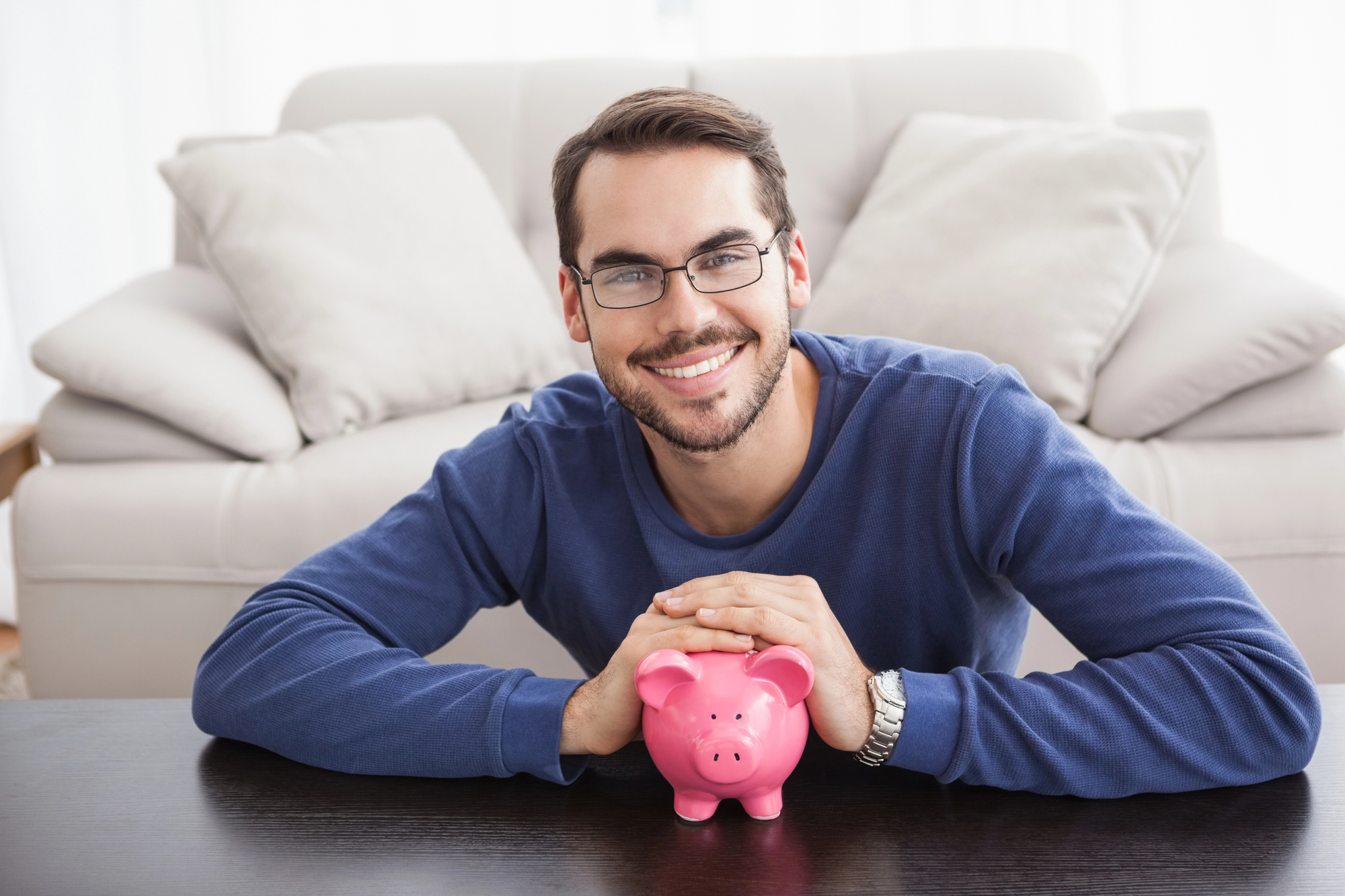 A smiling man with glasses and a beard, wearing a blue sweater, leans forward and rests his hands on a pink piggy bank placed on a dark wooden table. A white sofa and light-filled room are visible in the background.