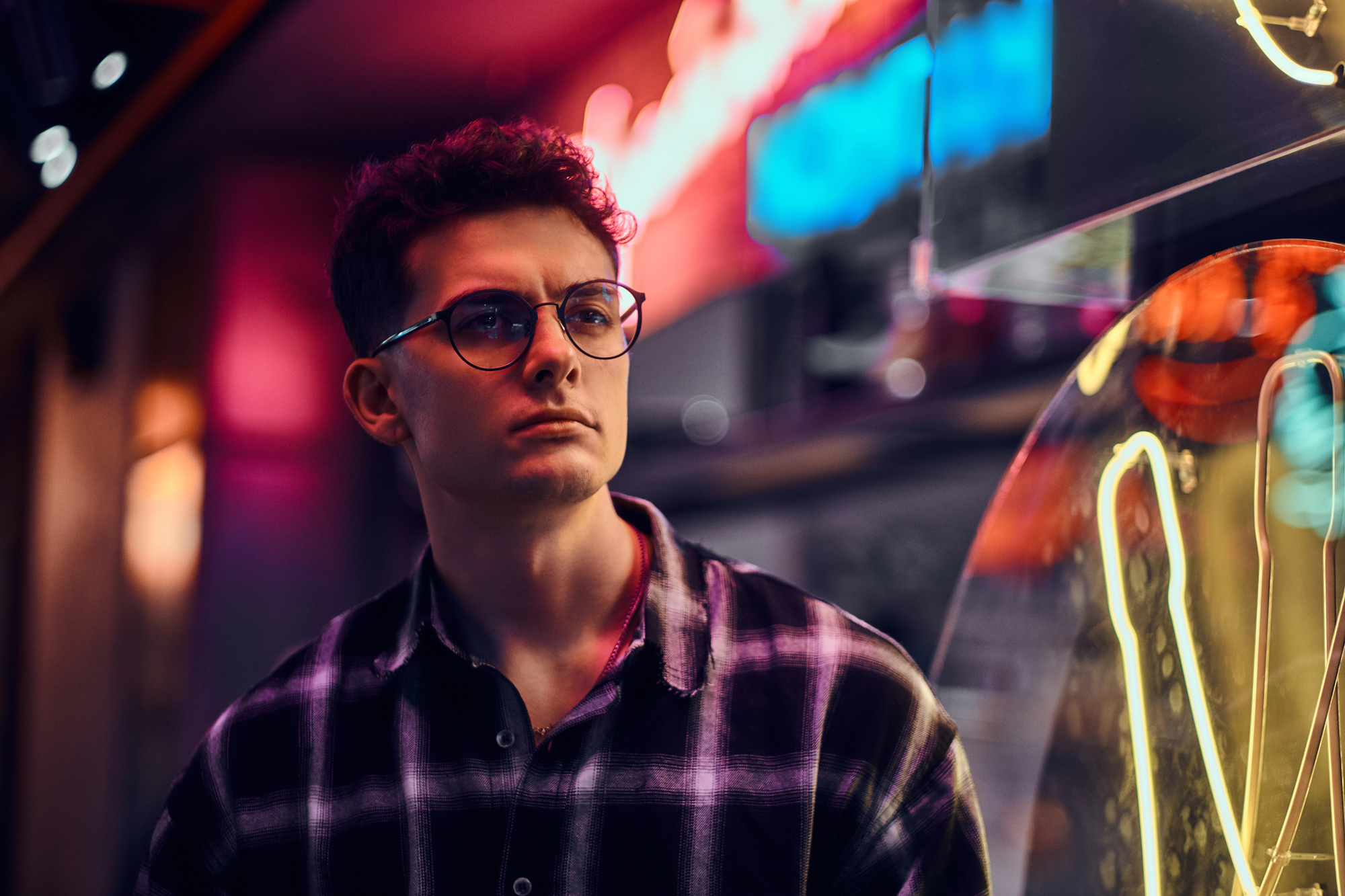 A young man in glasses and a checkered shirt stands in front of neon lights, gazing into the distance. The colorful lights create a vibrant and moody atmosphere, reflecting off the glass behind him.