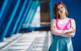 A woman with short red hair stands confidently against a blue wall. She wears a bright pink top, denim skirt, and matching pink sunglasses on her head. The background features a series of blue structural beams and patterns, with sunlight casting shadows.