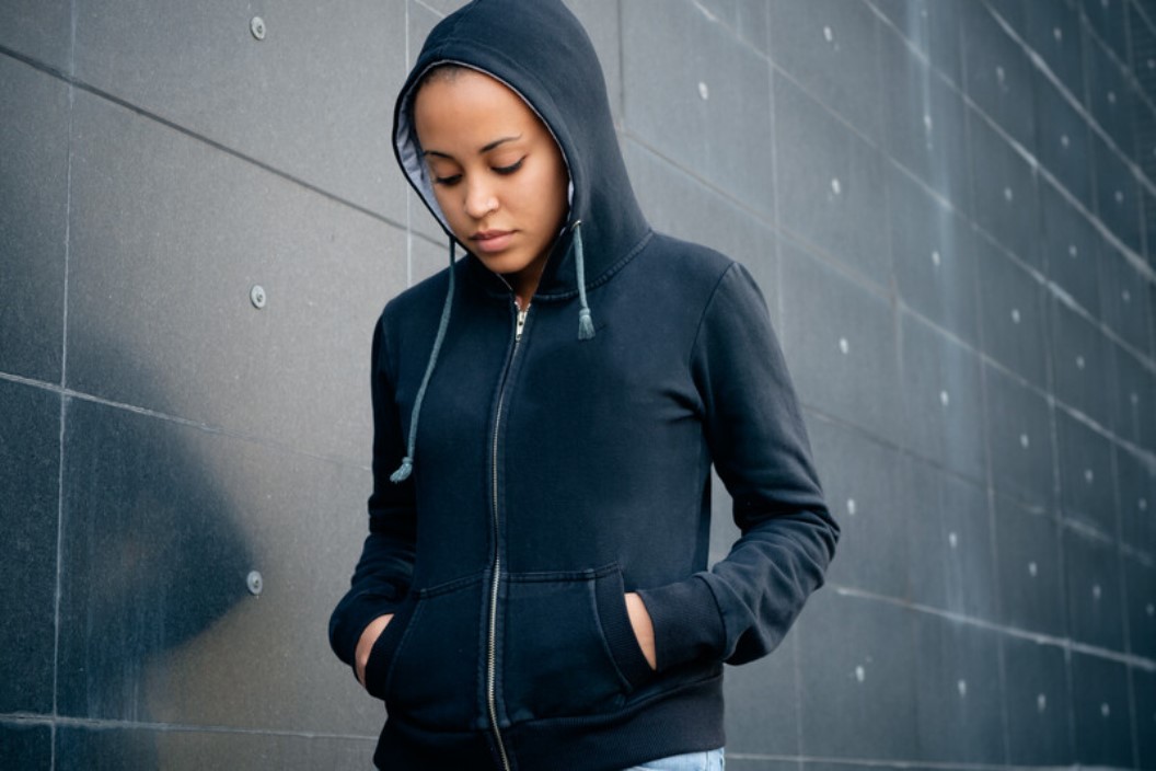 A person wearing a dark hoodie stands against a tiled wall with hands in the hoodie pockets. Their head is down, and the hood is up, partially shadowing their face, creating a contemplative or somber mood. The wall they lean against is dark and slightly reflective.