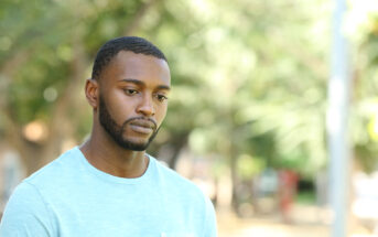 A young man with a short beard and wearing a light blue shirt stands outdoors, looking pensive with a slightly downcast gaze. The background is a blur of greenery and sunlight.