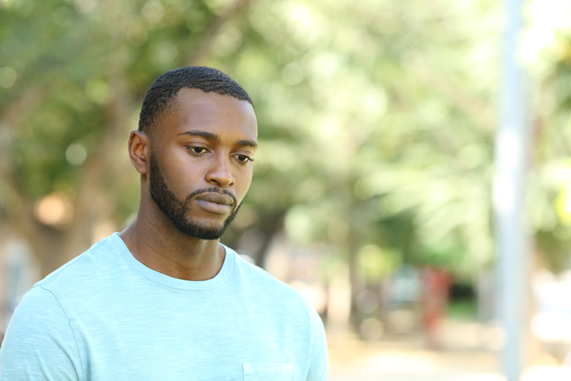 A young man with a short beard and wearing a light blue shirt stands outdoors, looking pensive with a slightly downcast gaze. The background is a blur of greenery and sunlight.