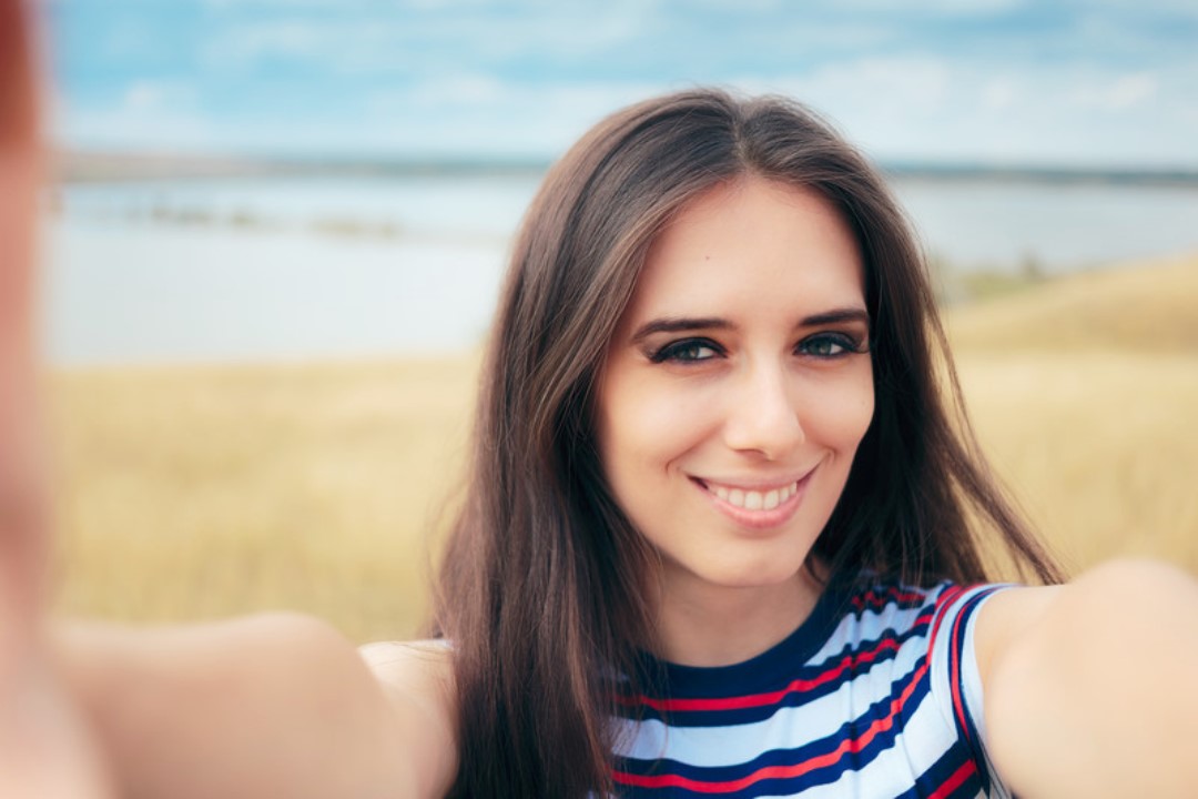 A young woman with long dark hair and a striped sleeveless shirt smiles at the camera, taking a selfie in an open field with a body of water in the background. The sky is partly cloudy.