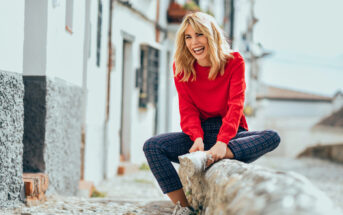 A woman wearing a red sweater and plaid pants is sitting on a stone wall in a narrow street with white buildings. She is smiling broadly and appears to be enjoying herself. The background is slightly blurred, highlighting her as the main subject.