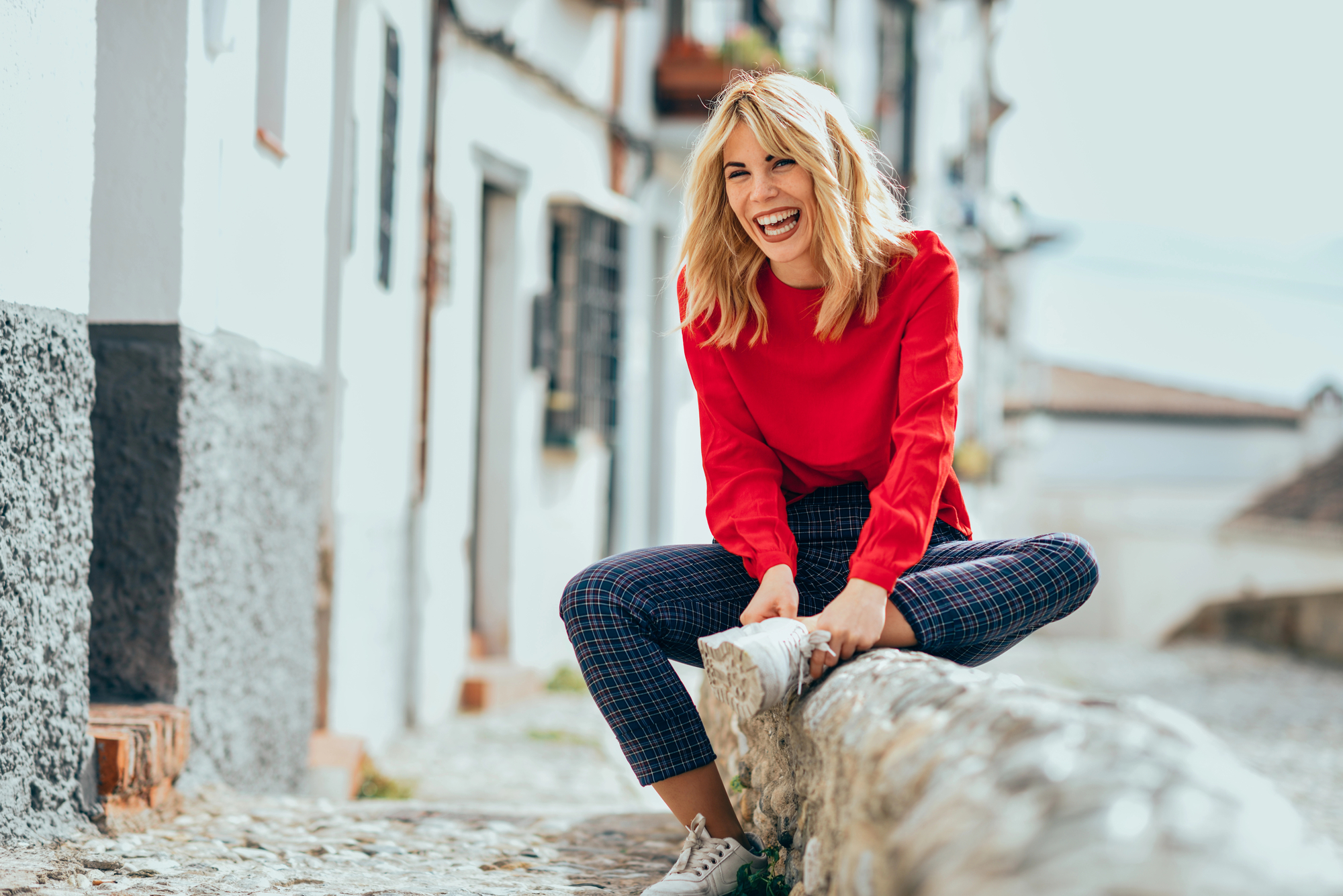 A woman wearing a red sweater and plaid pants is sitting on a stone wall in a narrow street with white buildings. She is smiling broadly and appears to be enjoying herself. The background is slightly blurred, highlighting her as the main subject.