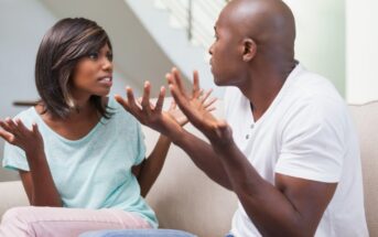 A man and a woman are sitting on a couch engaged in a heated discussion. The woman, wearing a light blue shirt, looks frustrated with her hands raised. The man, in a white t-shirt, also has his hands up, appearing to explain or argue back.