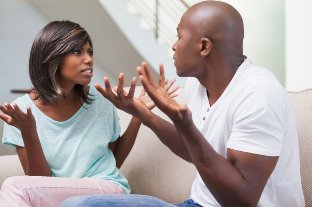 A man and a woman are sitting on a couch engaged in a heated discussion. The woman, wearing a light blue shirt, looks frustrated with her hands raised. The man, in a white t-shirt, also has his hands up, appearing to explain or argue back.