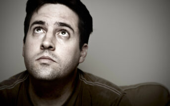 A man with a contemplative expression looks upward. He has short dark hair and a stubbly beard, and is wearing a brown shirt. The background is neutral and blurred, focusing attention on his face. The lighting is soft, casting gentle shadows.