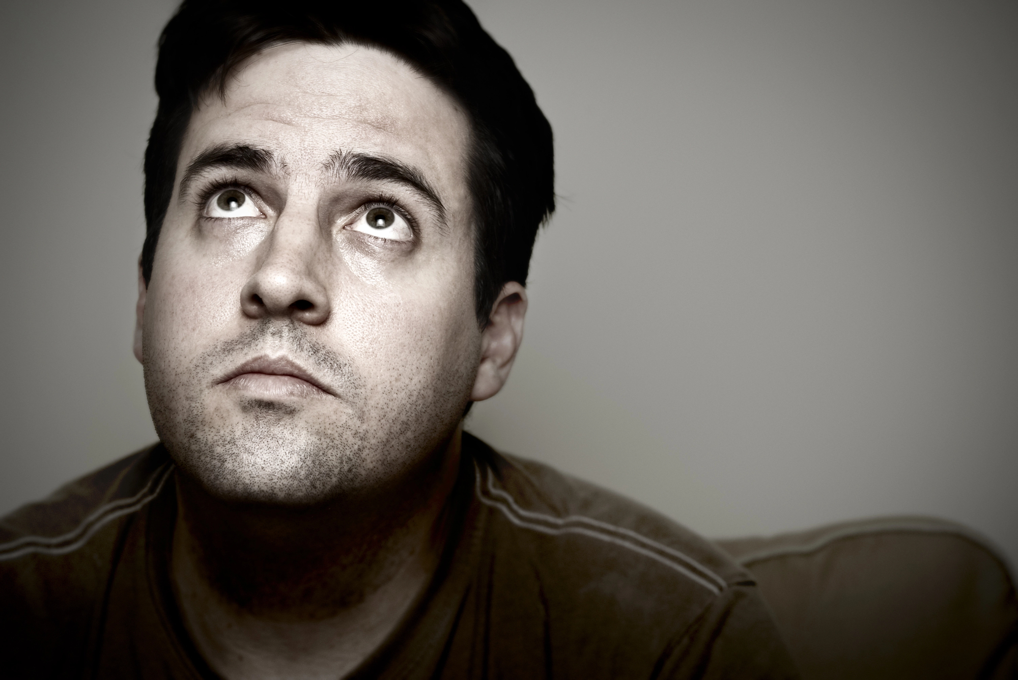 A man with a contemplative expression looks upward. He has short dark hair and a stubbly beard, and is wearing a brown shirt. The background is neutral and blurred, focusing attention on his face. The lighting is soft, casting gentle shadows.