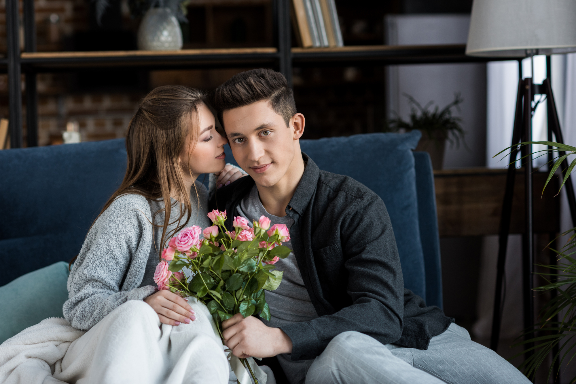 A woman and a man sit closely on a couch. The woman, wrapped in a blanket, is whispering into the man's ear while smiling. The man is holding a bouquet of pink roses and looking at the camera. They are in a cozy, modern living room with shelves and plants in the background.