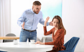 A man angrily shouts at a woman sitting at a white table. She looks distressed, raising one hand in a defensive gesture. Both have coffee cups in front of them. The setting appears to be a modern, minimalistic room with light-colored walls and furniture.