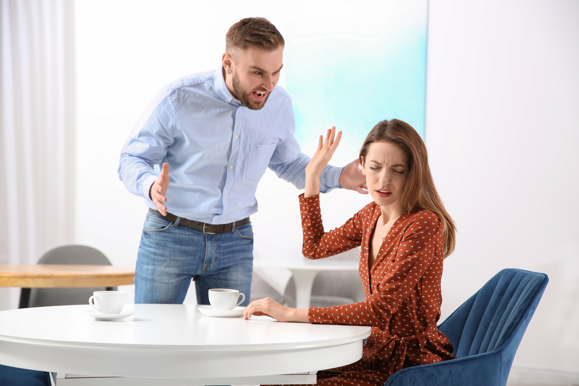 A man angrily shouts at a woman sitting at a white table. She looks distressed, raising one hand in a defensive gesture. Both have coffee cups in front of them. The setting appears to be a modern, minimalistic room with light-colored walls and furniture.