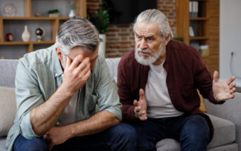 Two elderly men sit on a couch in a living room. One man, in a green shirt, leans forward with his head in his hand, appearing distressed. The other man, in a maroon shirt, gestures with his hands as if offering advice or support. Shelves and a TV are in the background.