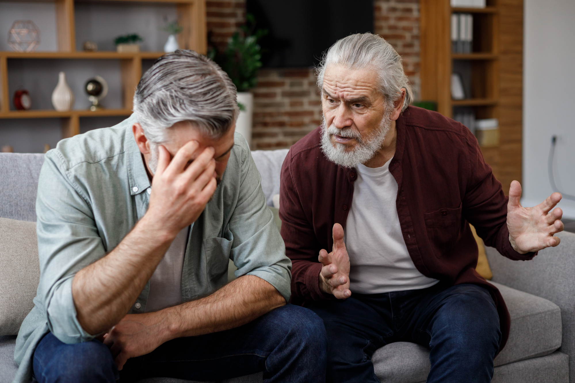 Two elderly men sit on a couch in a living room. One man, in a green shirt, leans forward with his head in his hand, appearing distressed. The other man, in a maroon shirt, gestures with his hands as if offering advice or support. Shelves and a TV are in the background.