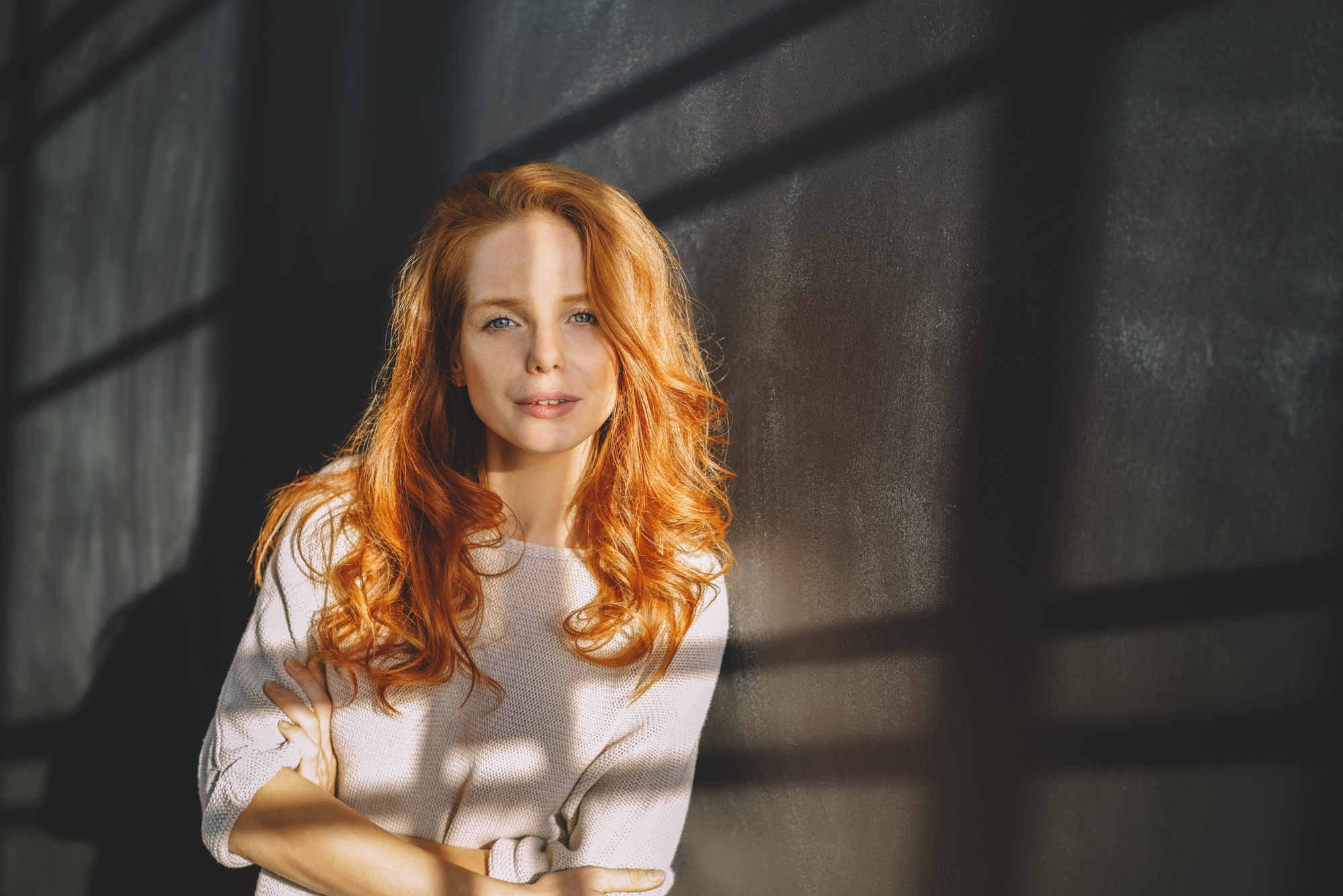 A woman with long, wavy red hair is standing against a dark wall with shadows cast from a window. She is wearing a light-colored sweater and has a relaxed expression, with her arms crossed, looking directly at the camera.