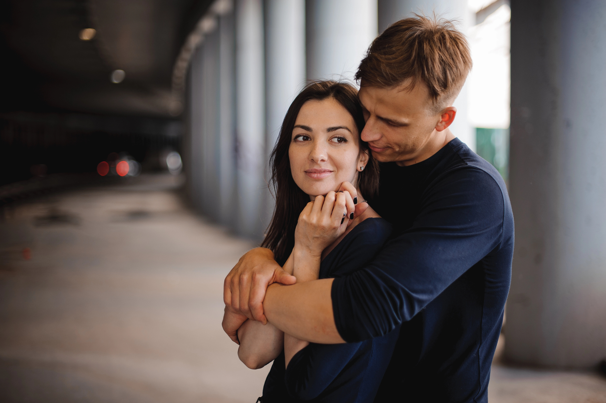 A young couple stands in a shaded area, with the man embracing the woman from behind. The woman smiles gently, holding his hands, while the man looks at her affectionately. Both are casually dressed in dark clothing, and the background is blurred with pillars.