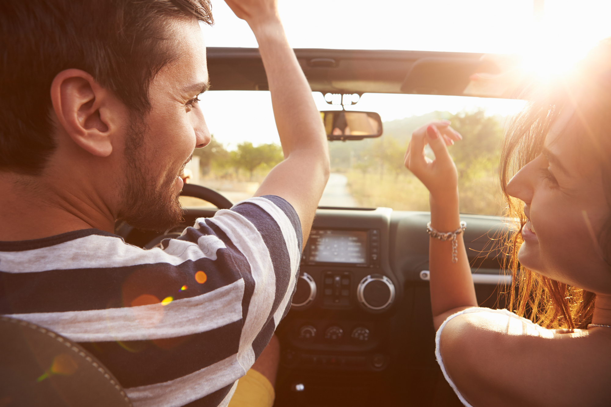 Two people are enjoying a drive in a convertible car. The sun is shining brightly, casting a warm glow on them. The person on the left, wearing a striped shirt, is driving, while the person on the right, with long hair, is smiling and raising their hand in joy.