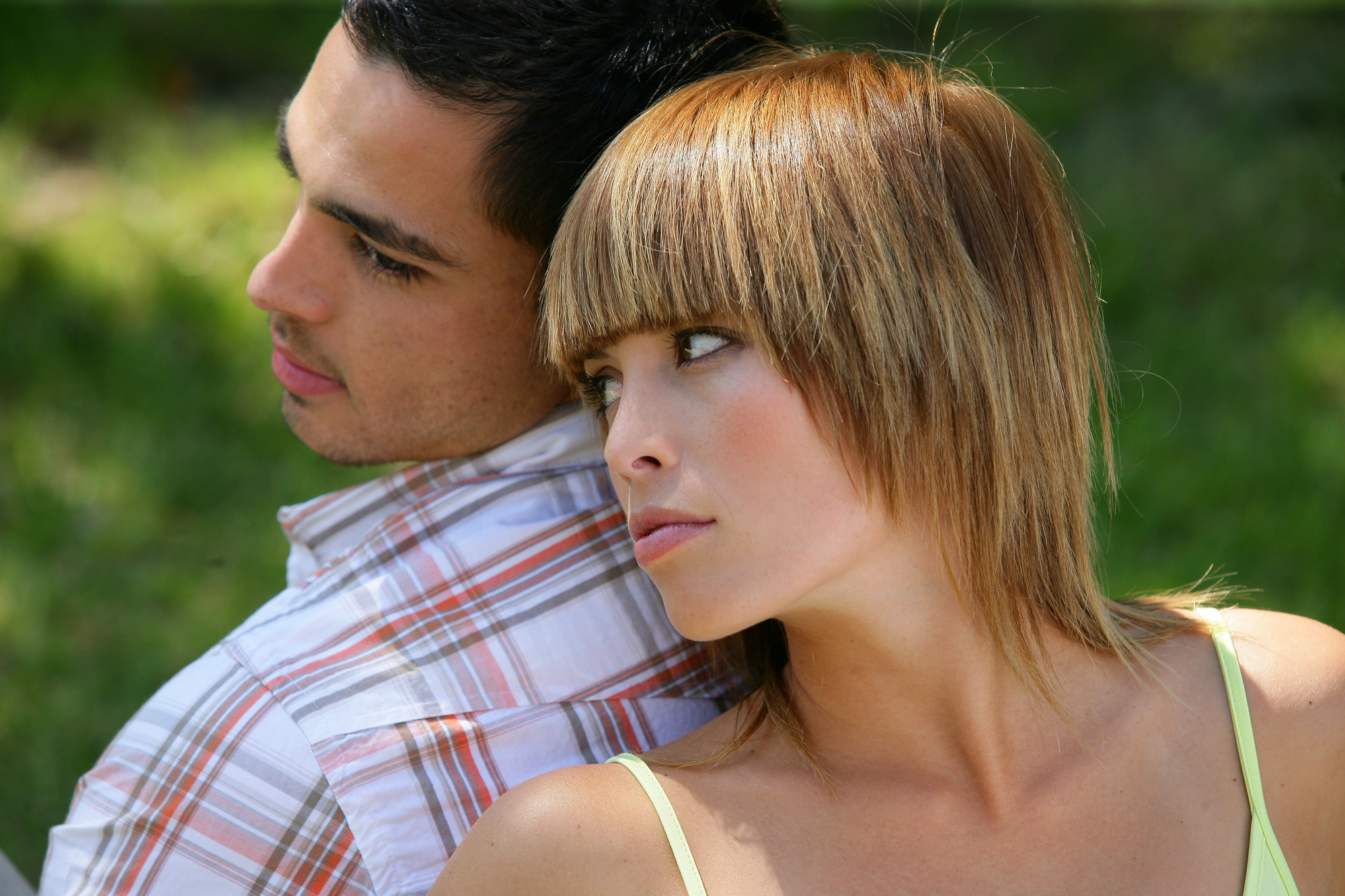 A man and a woman are sitting back-to-back in an outdoor setting. The man is wearing a plaid shirt and looking to the left. The woman, with short blonde hair and a sleeveless top, is looking to the right with a slightly serious expression. Both appear deep in thought.