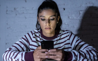 A woman with long dark hair, wearing a striped shirt, sits at a table in front of a white brick wall, focused on her smartphone. The lighting is dim, casting shadows on the wall behind her.