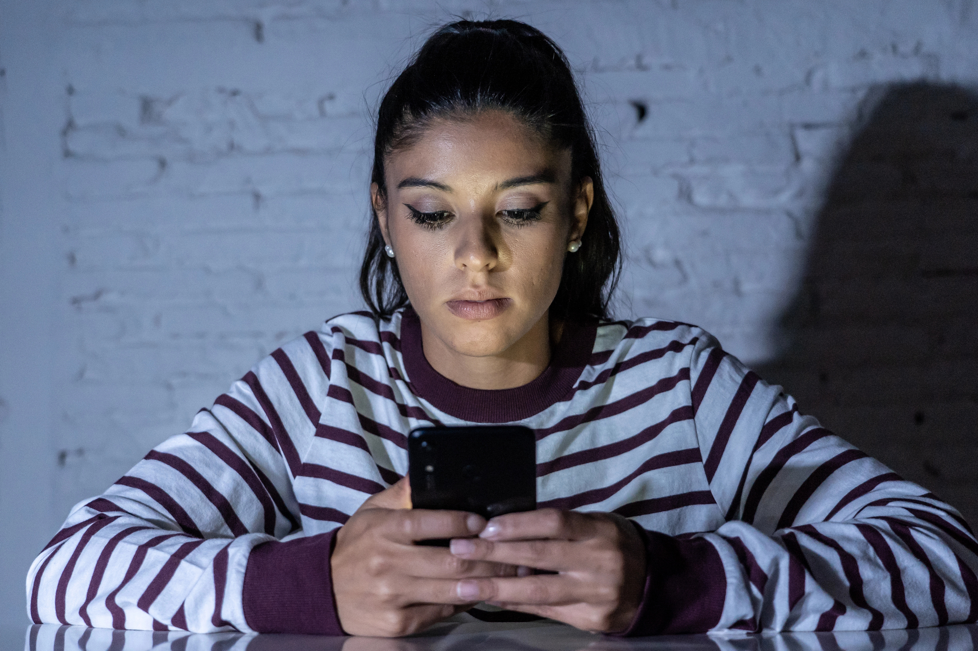 A woman with long dark hair, wearing a striped shirt, sits at a table in front of a white brick wall, focused on her smartphone. The lighting is dim, casting shadows on the wall behind her.