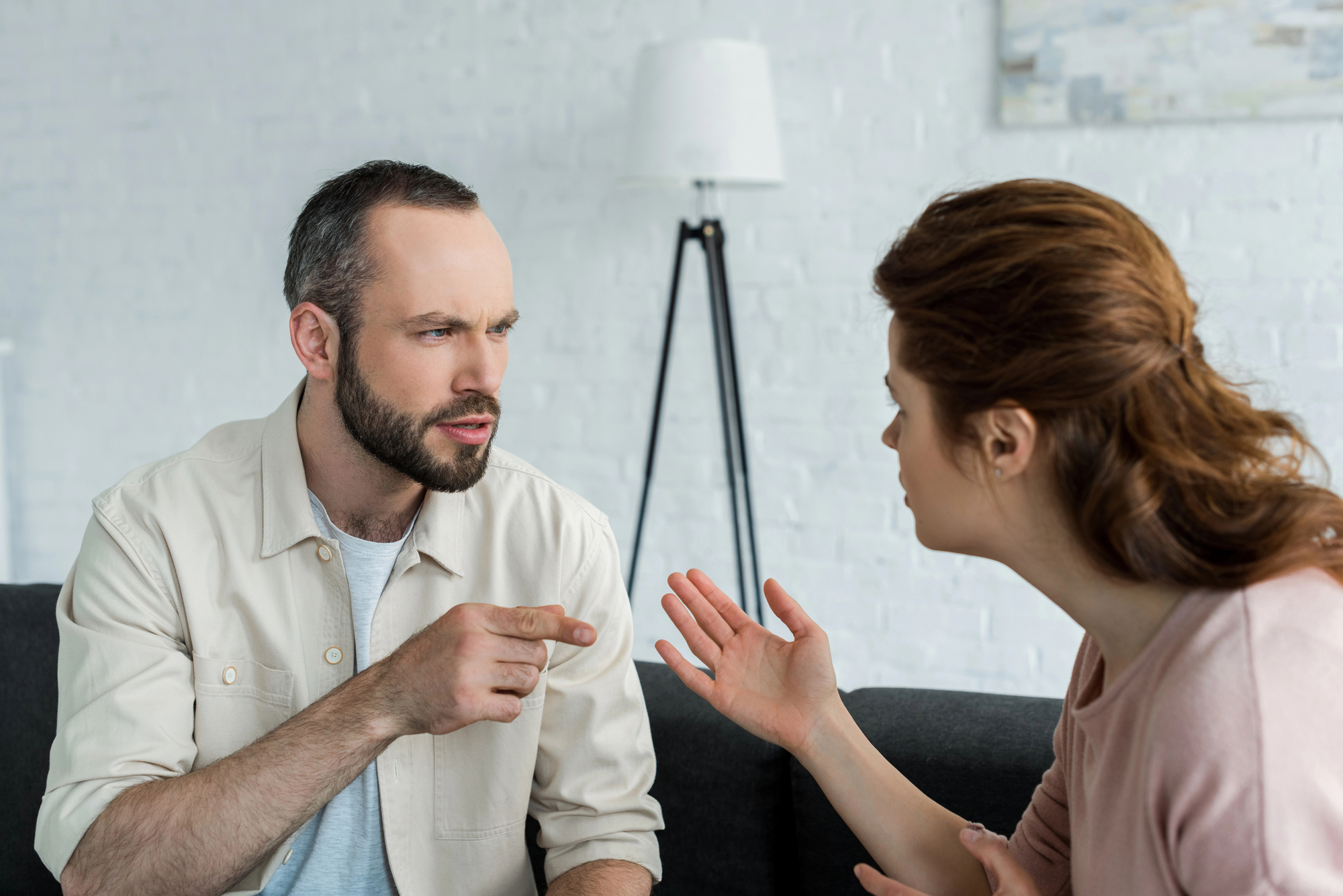 A man and a woman are having a serious conversation on a couch. The man, with a serious expression, is gesturing with his finger, while the woman is gesturing with her hands, appearing to explain or argue. A lamp and a textured white wall are in the background.