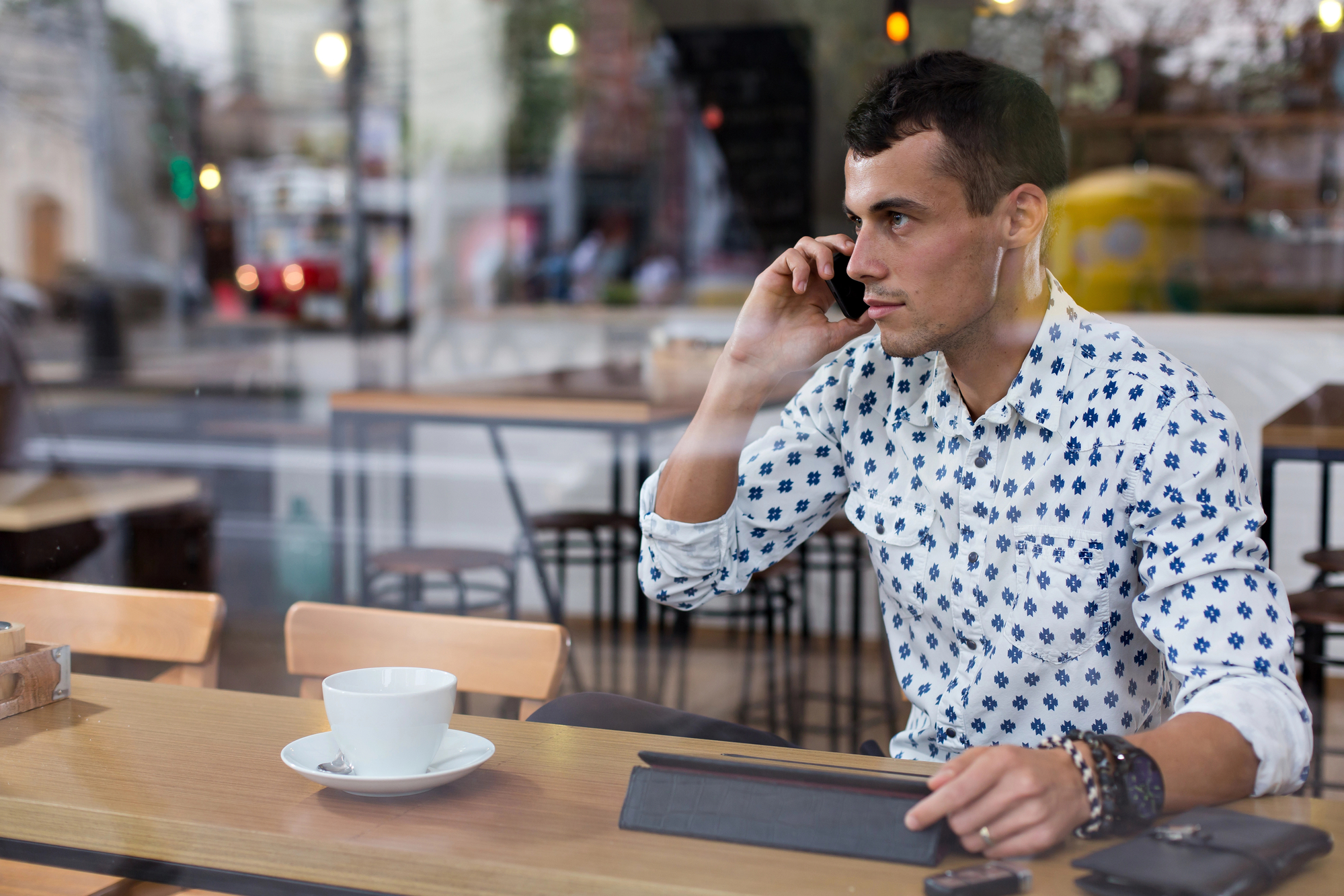 A man with short dark hair sits at a wooden table in a cafe, talking on his smartphone. He is wearing a white shirt with a blue pattern and several bracelets on his wrist. A white coffee cup and saucer are in front of him, and the street is visible through the window.