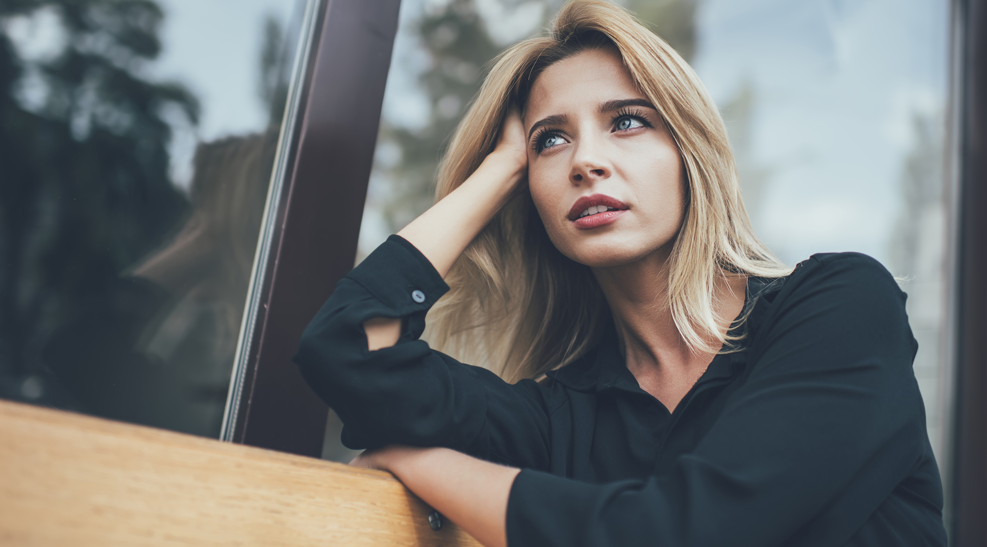 A woman with long blonde hair, wearing a black shirt, is sitting on a wooden bench. She has one hand resting on her head and is looking thoughtfully into the distance. The background is blurred with reflections on a glass surface behind her.