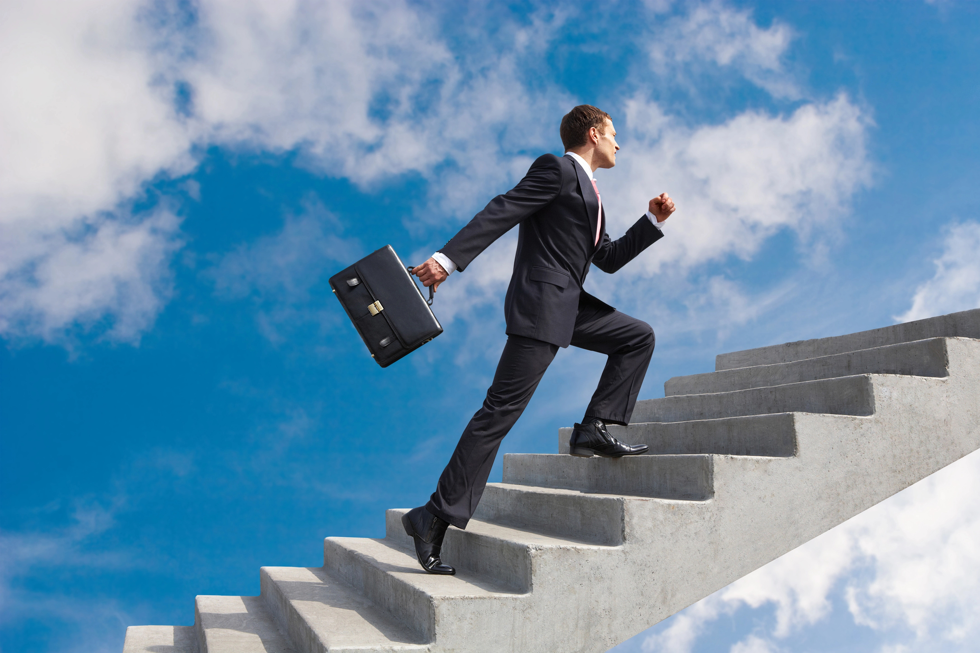 A man in a suit holds a briefcase and ascends an outdoor staircase that appears to climb into the sky. The scene conveys ambition and progress, set against a backdrop of a bright blue sky with scattered clouds.