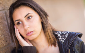 A young woman with shoulder-length brown hair leans against a wall, resting her cheek on her hand. She wears a dark jacket with a leopard print lining. She has a thoughtful or slightly pensive expression on her face.