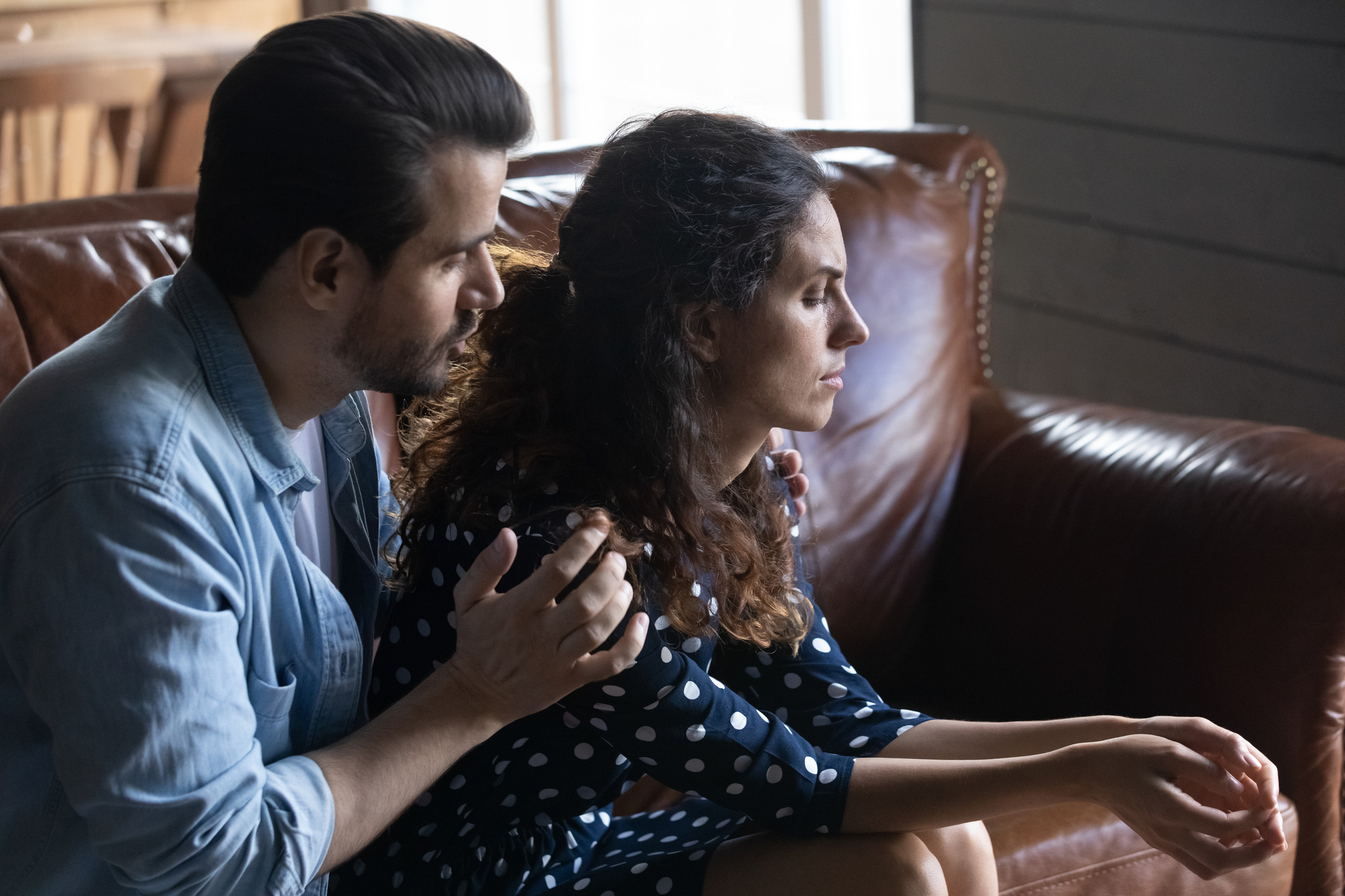 A man sits beside a woman on a brown leather couch, gently placing his hand on her shoulder in a comforting manner. The woman looks pensive, staring ahead, dressed in a polka dot dress. The scene is set in a cozy room with a wooden background.