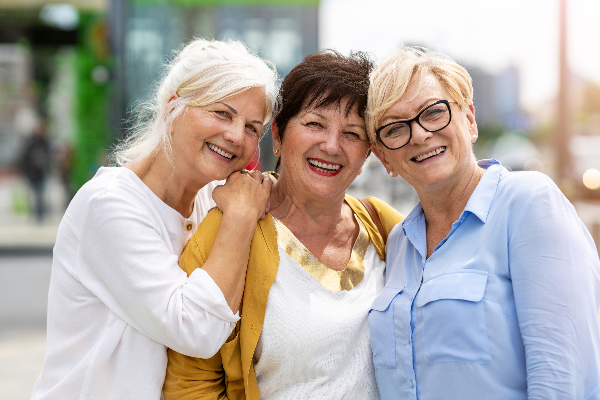 Three senior women smiling and posing together outdoors. The woman on the left has white hair and is wearing a yellow jacket, the woman in the middle has dark hair and a white top, and the woman on the right has blond hair, glasses, and a blue blouse.