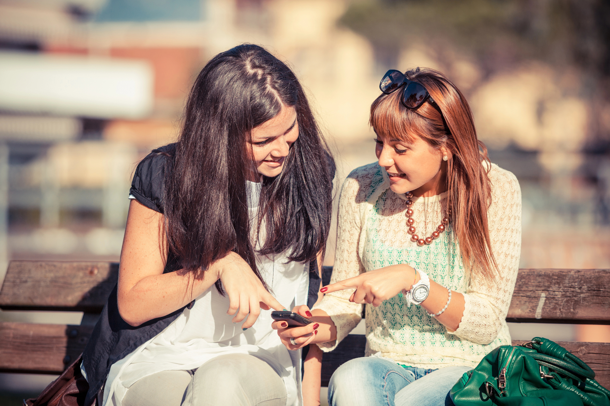 Two young women sitting on a bench outdoors, both smiling and looking at a smartphone held by the woman with dark hair. The woman with lighter hair is pointing at the phone. The background appears to be a sunny day with blurred buildings and trees.