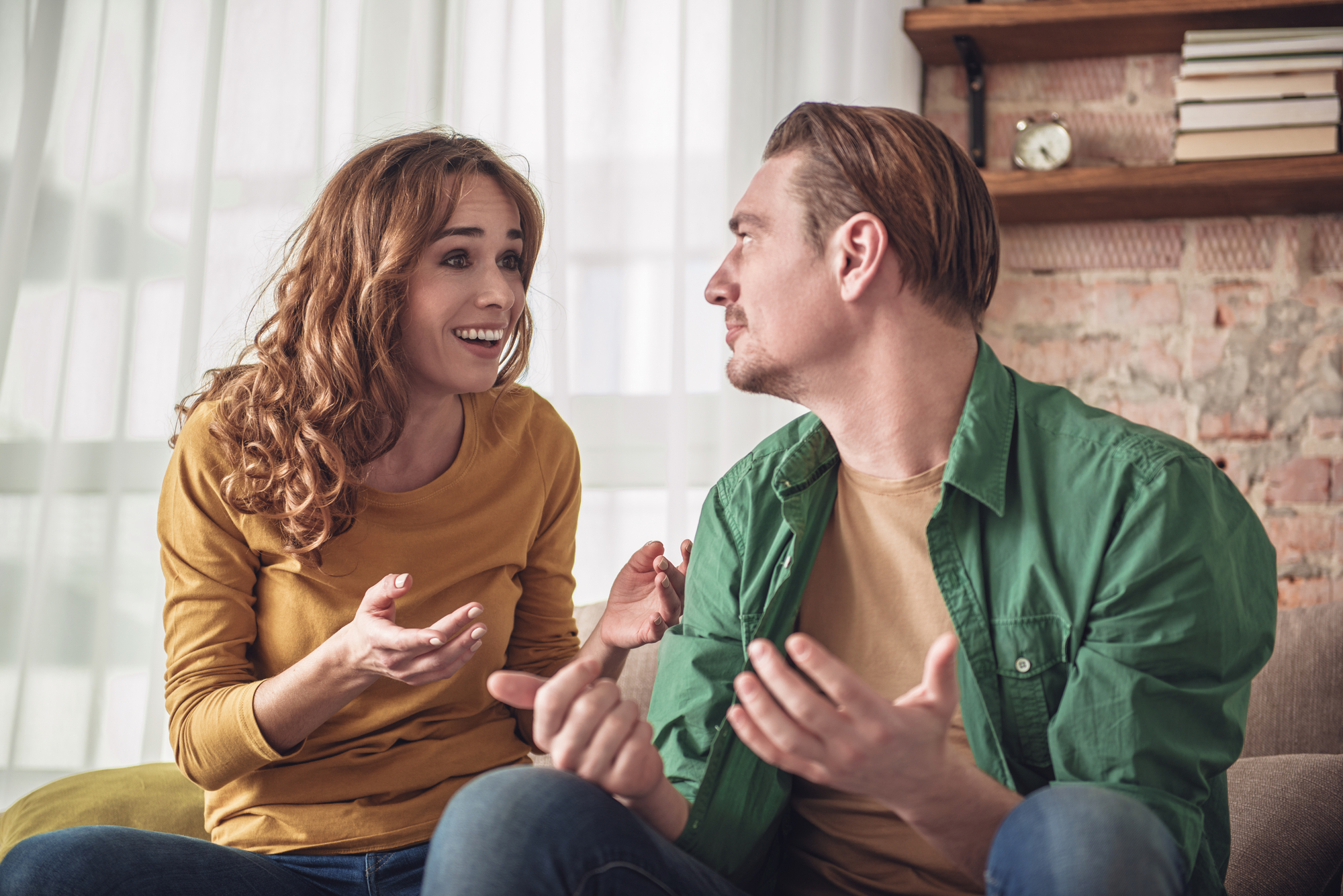 A man and woman are sitting on a couch having a discussion. The woman, with curly hair and wearing a yellow top, has an animated expression with her hands gesturing. The man, with short hair and wearing a green shirt, is facing her and seemingly responding.