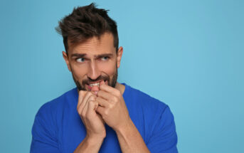 A man with short brown hair and a beard, wearing a blue shirt, stands in front of a blue background, appearing anxious and nervous with hands near his mouth, biting his nails. He has an uneasy expression, looking slightly to his right.