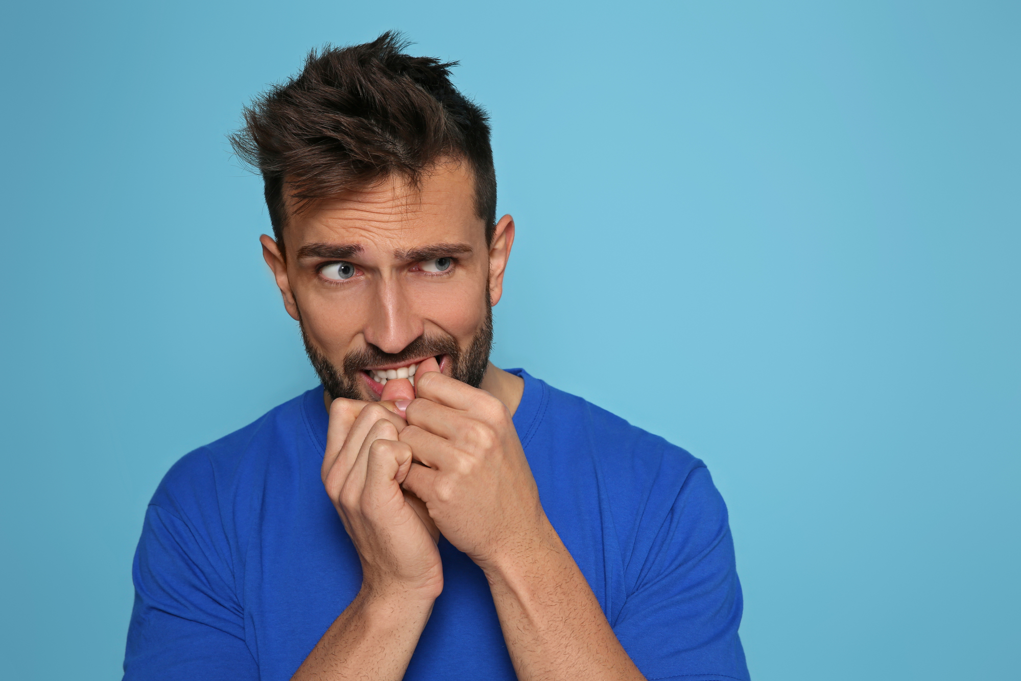 A man with short brown hair and a beard, wearing a blue shirt, stands in front of a blue background, appearing anxious and nervous with hands near his mouth, biting his nails. He has an uneasy expression, looking slightly to his right.