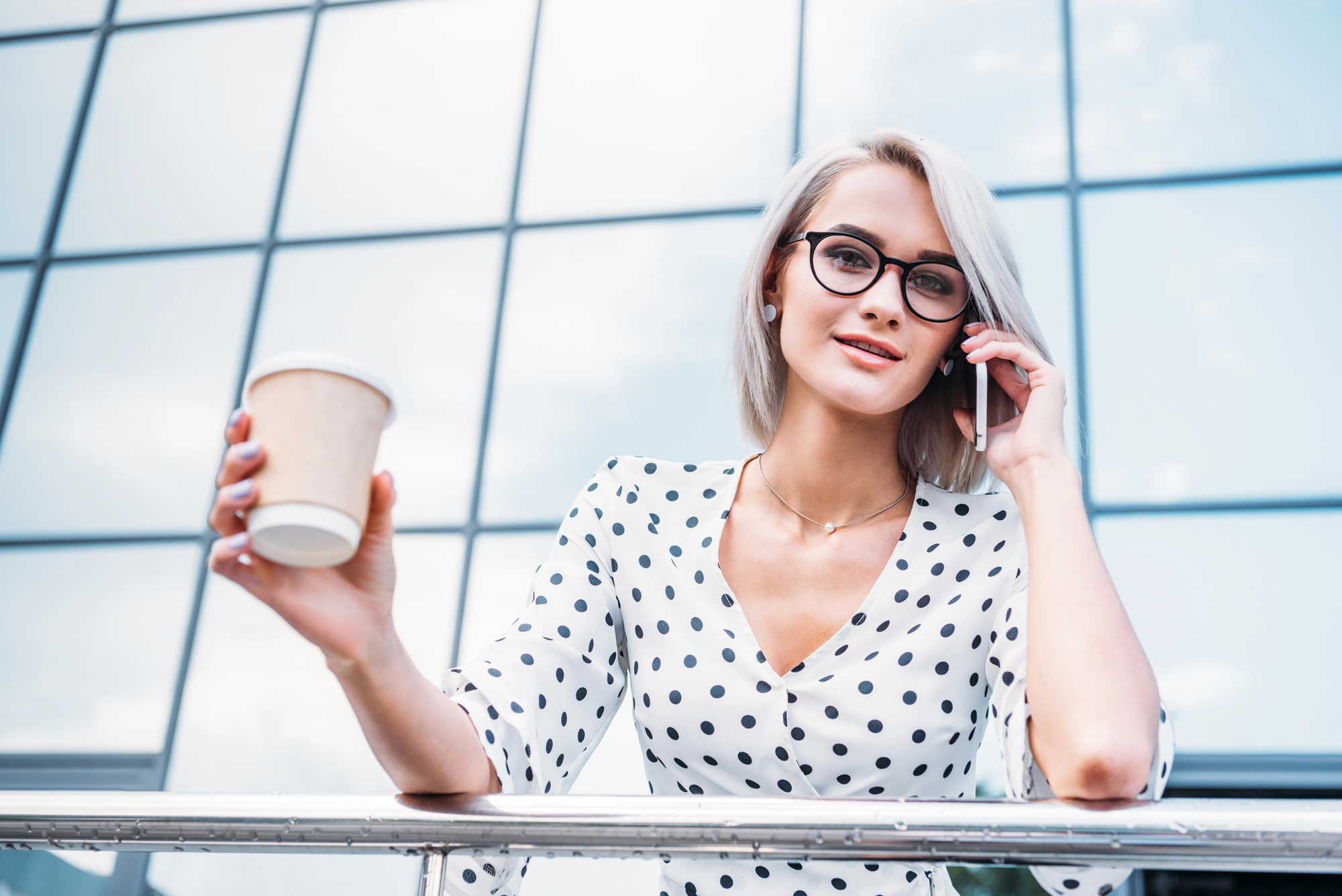 A person with short blond hair, wearing glasses and a white polka dot blouse, is smiling while talking on a mobile phone. They are holding a takeaway coffee cup and standing in front of a glass building facade.