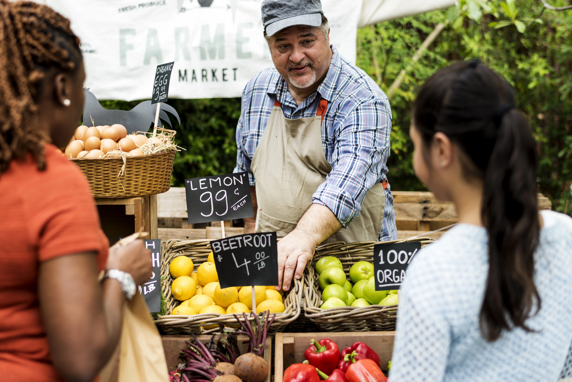 A man in an apron stands behind a market stall, handing fresh produce to a customer. The stall displays baskets of lemons, limes, beetroot, eggs, and other vegetables, with small signs showing prices. Two women stand in front of the stall, interacting with the seller.