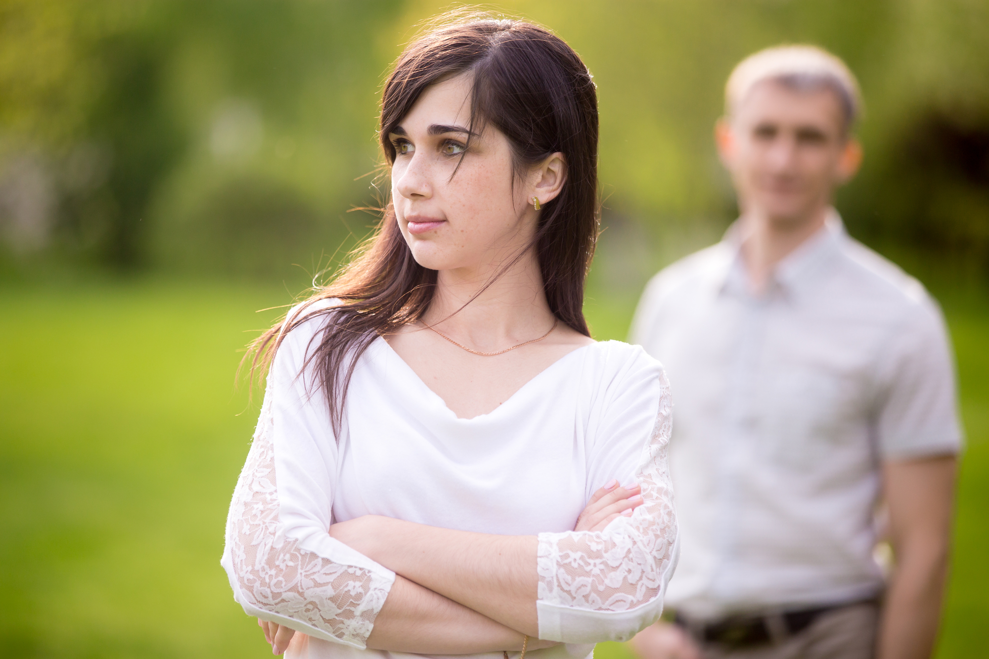 A young woman with long dark hair stands outdoors with her arms crossed, looking to the side. She is wearing a white top with lace sleeves. A man, slightly out of focus, stands in the background wearing a light-colored shirt, smiling gently. Greenery surrounds them.