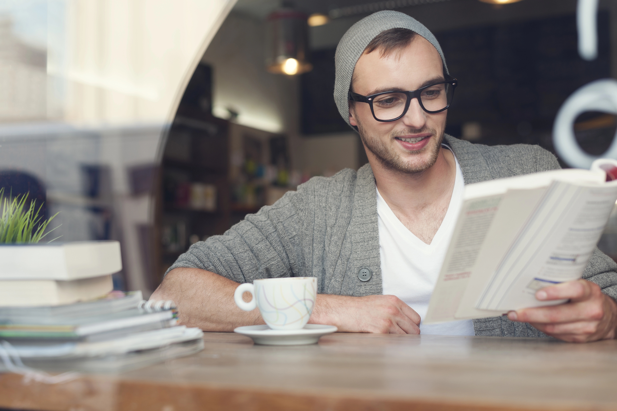 A man wearing glasses and a gray beanie is sitting at a wooden table in a cozy cafe. He is reading a book and smiling. A cup of coffee, a stack of books, a potted plant, and a notebook with a pen are on the table. The background is softly lit.