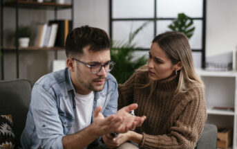 A woman with long hair in a brown sweater sits closely beside a man with short dark hair, glasses, and a denim shirt, who appears distressed. She comforts him with a concerned expression as he gestures with his hands, sitting on the couch in a cozy room.