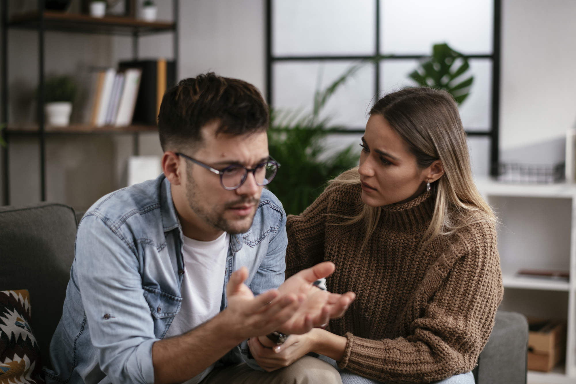 A woman with long hair in a brown sweater sits closely beside a man with short dark hair, glasses, and a denim shirt, who appears distressed. She comforts him with a concerned expression as he gestures with his hands, sitting on the couch in a cozy room.