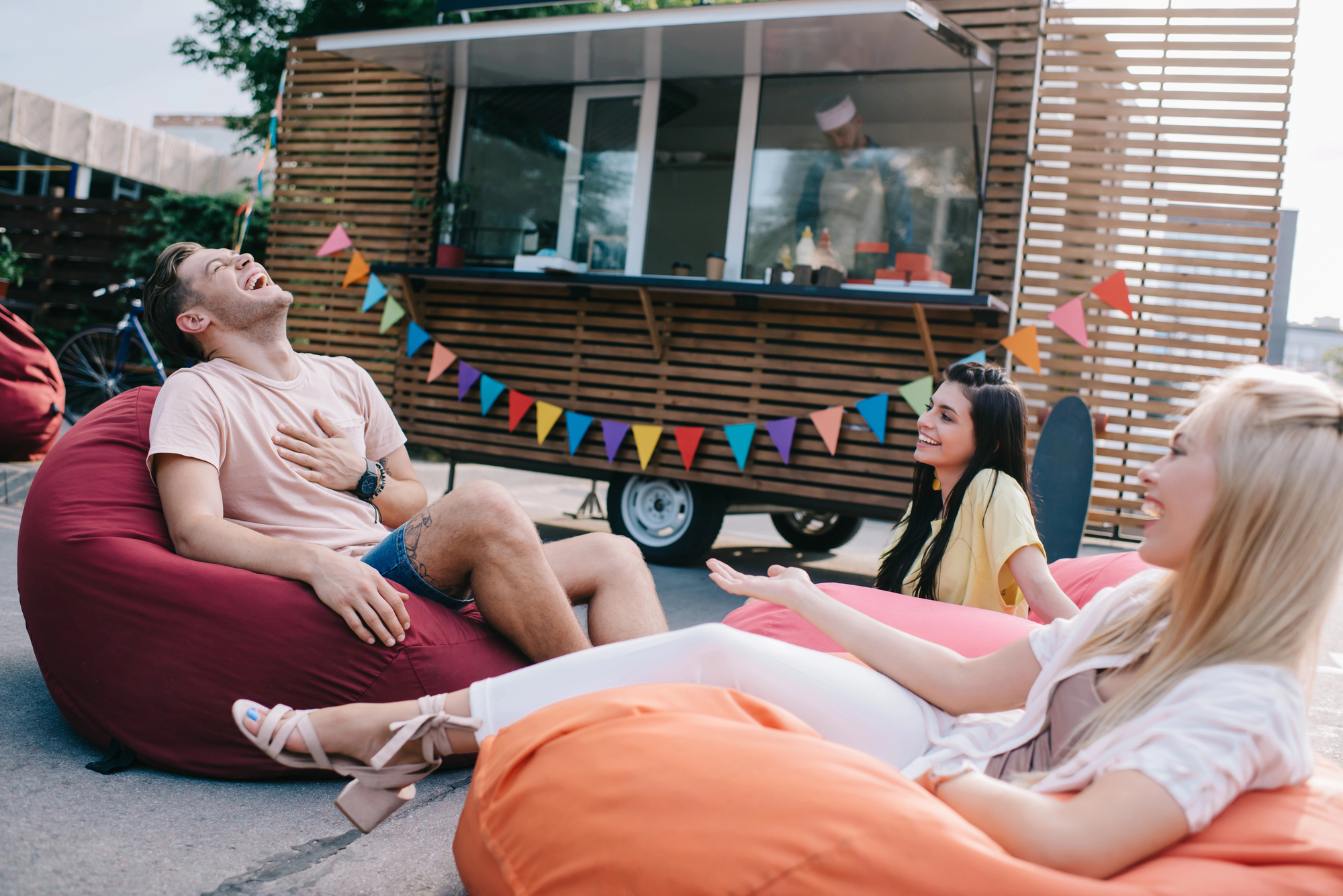 Three people are relaxing on colorful bean bags outdoors, laughing and chatting. Behind them is a food truck with a wooden facade and colorful triangular bunting. The setting appears casual and cheerful, perfect for a friendly gathering.