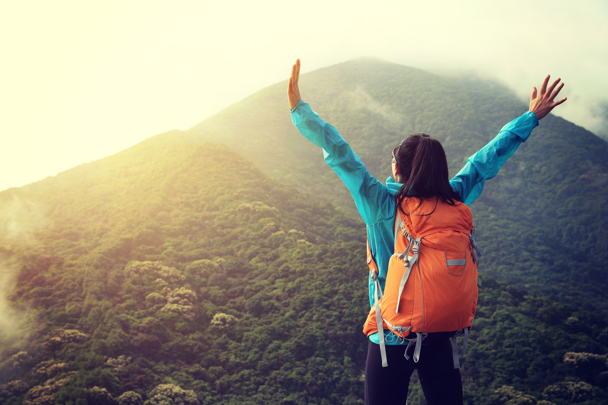 A person wearing a bright blue jacket and an orange backpack stands on a mountain summit with arms raised in triumph, overlooking a lush, green landscape shrouded in mist. The morning sun breaks through clouds, illuminating the scene with a warm, golden light.