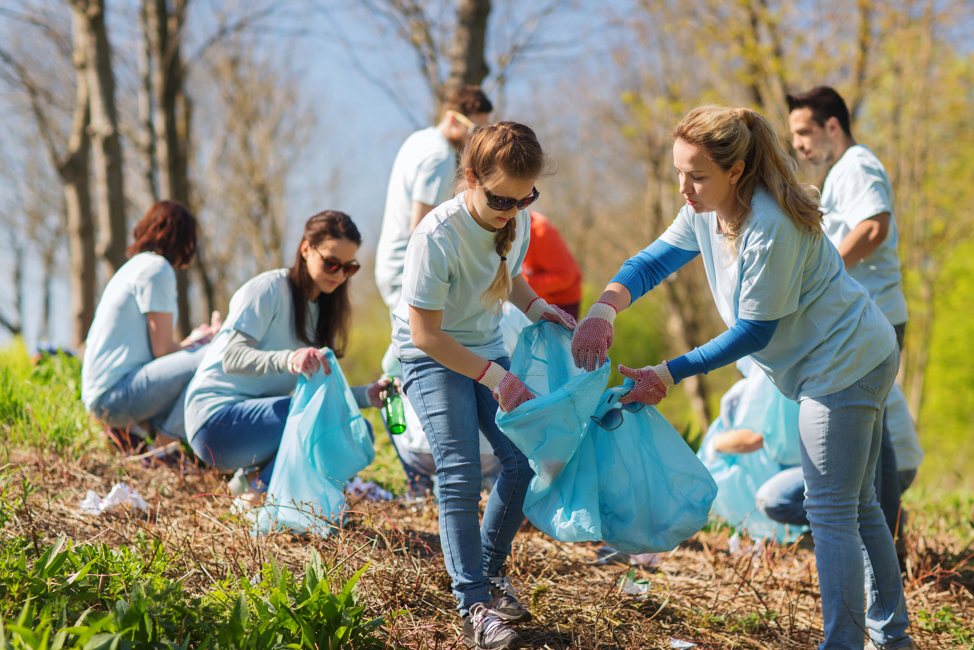 A group of people wearing light blue shirts and gloves are participating in an outdoor clean-up activity. They are collecting trash and debris in blue plastic bags on a grassy area with trees in the background under a sunny sky.