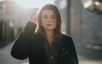 A young woman with long brown hair stands outdoors in a sunlit, urban setting. She is wearing a dark coat and looking directly at the camera, with one hand raised to her hair. The background is slightly blurred, showing buildings in soft focus.