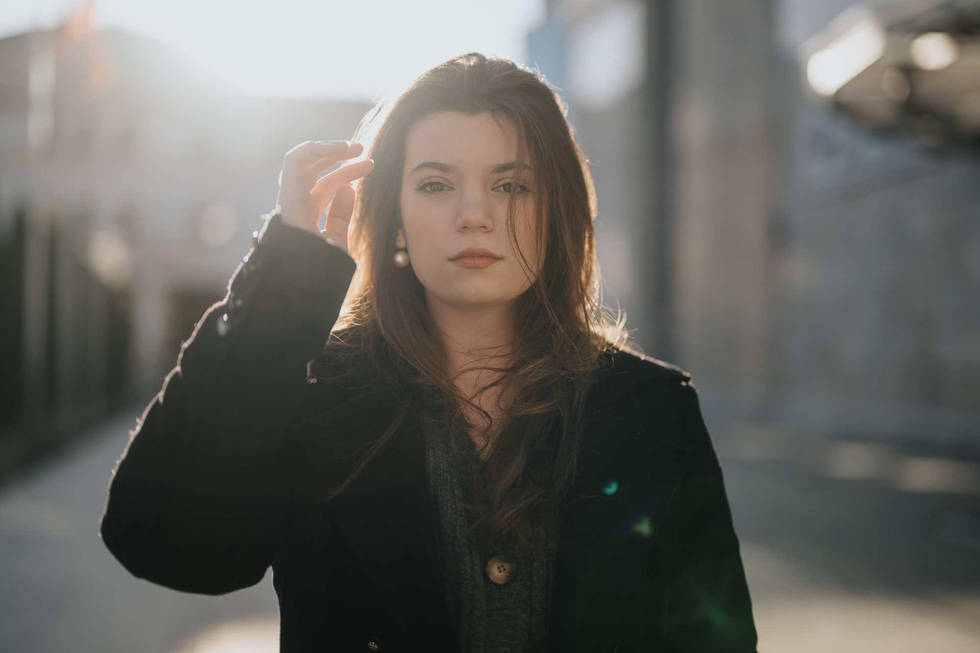 A young woman with long brown hair stands outdoors in a sunlit, urban setting. She is wearing a dark coat and looking directly at the camera, with one hand raised to her hair. The background is slightly blurred, showing buildings in soft focus.