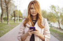 A woman with long red hair is standing on a paved path in a park, holding a purple smartphone in both hands. She is wearing a light-colored jacket and looking directly at the camera. Trees and greenery are visible in the background.